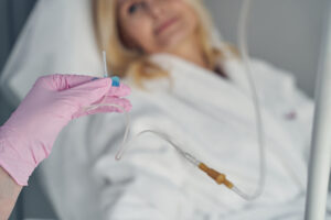 Gloved hand holding a IV needle with a woman looking on in the background